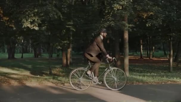 Молодий чоловік їде на велосипеді в міському парку в діловому костюмі і шоломі, вид збоку — стокове відео