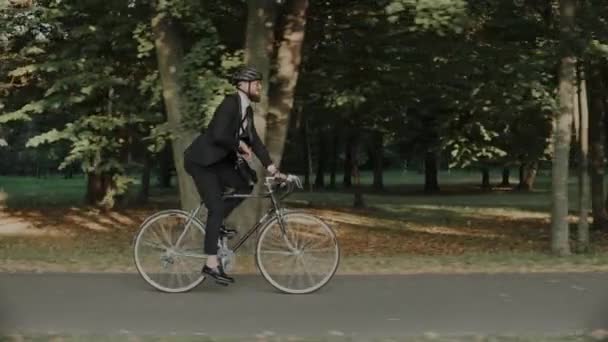 Hombre de negocios en bicicleta en el parque de la ciudad con casco y una bolsa, vista lateral — Vídeos de Stock