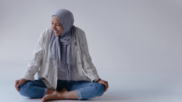 Młoda muzułmanka siedzi w jasnobiałym studio uśmiechnięta i śmiejąca się z kamery — Wideo stockowe