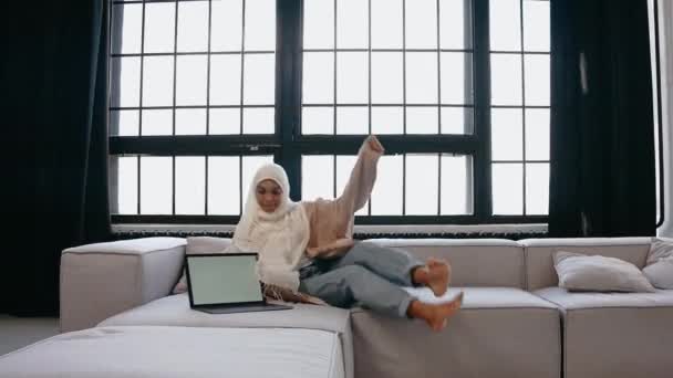 Ung muslim flicka sitter på soffan och tittar på laptop i ljusa beige studio — Stockvideo