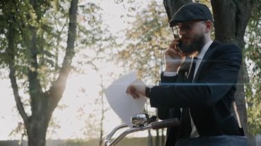 İş adamı telefonda konuşuyor, saatleri kontrol ediyor, şapka ve gözlük takıyor.