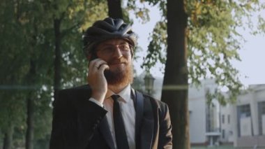 Bisiklet kaskı takan bir iş adamı telefonda konuşurken parkta gülümsüyor.