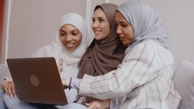 Üç Müslüman kız dizüstü bilgisayara bakar, gülümser ve parlak bej renkli stüdyoda güler.