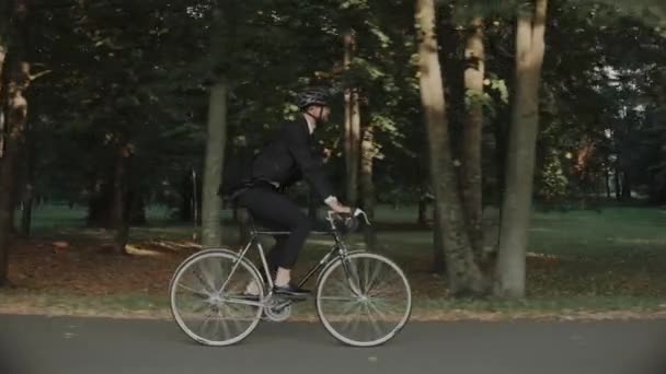 Empresário montando uma bicicleta no parque da cidade usando capacete e um saco, vista lateral — Vídeo de Stock