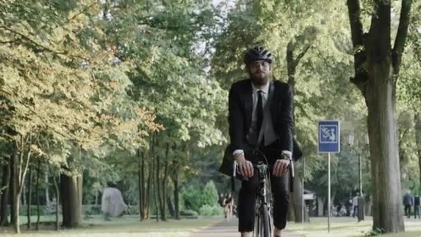 Empresário montando uma bicicleta no parque da cidade vestindo terno e capacete, vista frontal — Vídeo de Stock