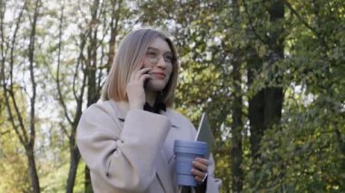 Genç bir kız telefonda konuşur ve güneşli bir sonbahar gününde palto giyerek gülümser.