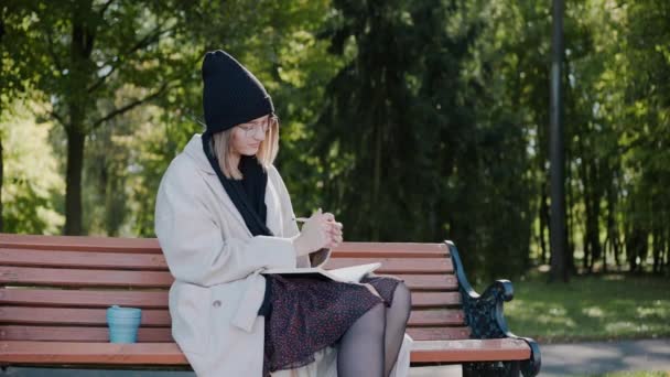 Jovem sentada em um banco de parque vestindo casaco e chapéu, escrevendo em caderno — Vídeo de Stock