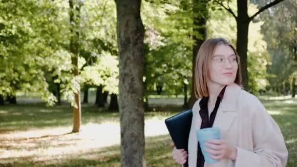 Молодая девушка гуляет в городском парке в солнечный осенний день в пальто и улыбается — стоковое видео