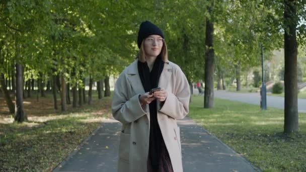 Menina caminha no parque da cidade em um dia ensolarado de outono mensagens de texto e sorrindo — Vídeo de Stock