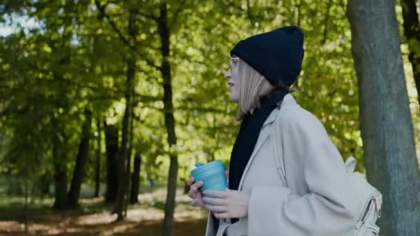 Chica camina en el parque de la ciudad en un día soleado de otoño, saludando a un amigo y sonriendo — Vídeos de Stock