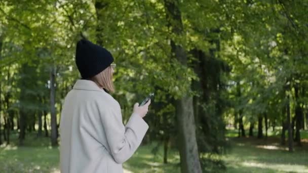 Güneşli bir sonbahar gününde şehir parkında yürüyen ve telefona bakan bir kız. — Stok video