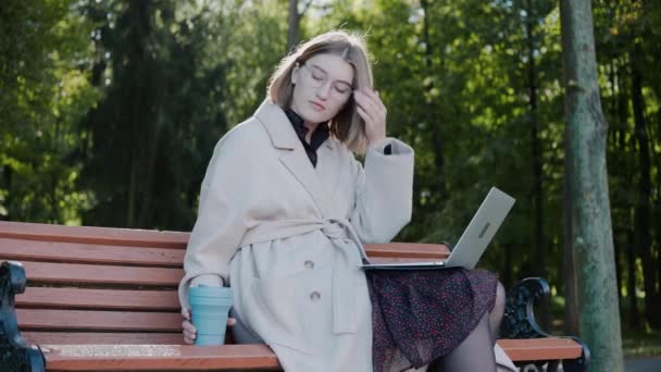 Ragazza si siede su una panchina in autunno, scrivendo su un computer portatile, utilizzando un telefono e bevendo — Video Stock
