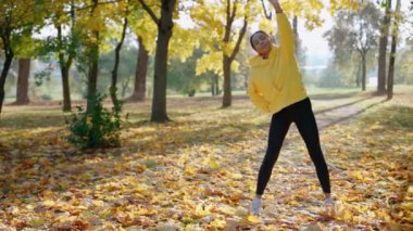 Kız sonbaharda parkta sabah koşusundan önce egzersiz ve esneme hareketleri yapıyor.