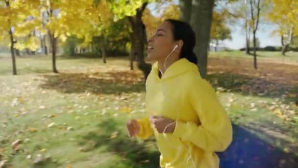 Menina correndo na pista no parque da cidade na manhã de outono ensolarada, usando fones de ouvido — Vídeo de Stock