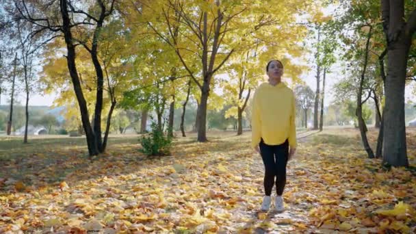 Дівчина робить стрибаючий джек і розтягується перед ранковим бігом в парку восени — стокове відео