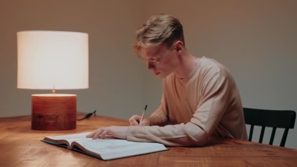Jovem estudando em casa escrevendo em suas anotações, lendo um livro na mesa — Vídeo de Stock