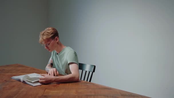 Szemüveges fickó tankönyvből tanul, üres szürke szobában ül. — Stock videók