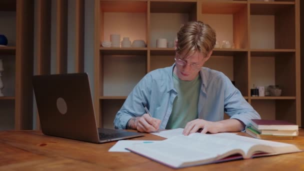 Парень, занимающийся дома, смотрит на ноутбук, пишет блокнот и читает учебник — стоковое видео