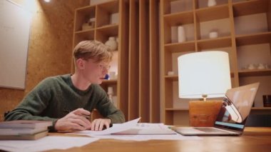 Genç adam evde ders çalışıyor, bilgisayarında bir video görüşmesi yapıyor, notlarını yazıyor.