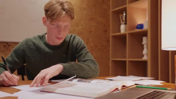 Jongeman die thuis studeert, naar de laptop kijkt, in een notitieboekje schrijft — Stockvideo