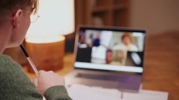 Joven estudiando en casa, haciendo una videollamada en un portátil, escribiendo sus notas — Vídeo de stock