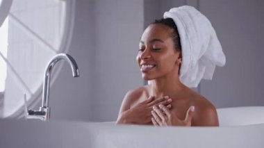 Banyo küvetinde boynunu ve omuzlarını yıkayan siyah kadın kameraya bakıyor.
