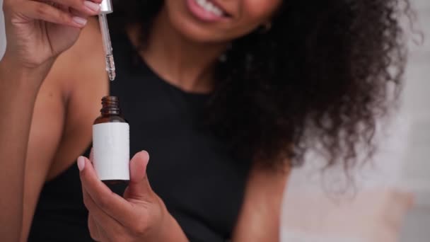 Mujer negra aplica aceite cosmético de una botella con gotero, sentado en una cama — Vídeos de Stock
