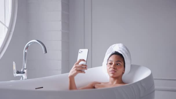 Donna nera sdraiata in un bagno a farsi selfie con i capelli avvolti in un asciugamano — Video Stock