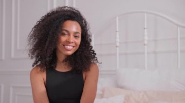 Siyah kadın saçlarını elleriyle sallıyor ve yatağa oturmuş kameraya bakıyor.