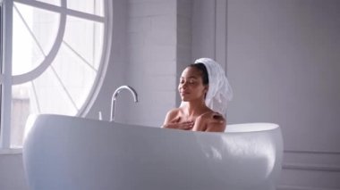 Siyah bir kadın havluya sarılmış saçlarıyla banyo yapıyor, yavaşça aşağı kayıyor.