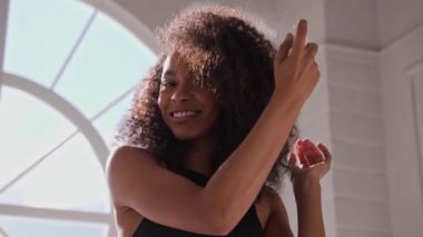 Siyah kadın saç spreyi sıkıyor, gülümsüyor ve kameraya bakıyor.