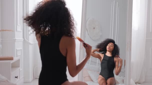 Μαύρη γυναίκα χτενίζει τα μαλλιά της, κάθεται στο κρεβάτι και κοιτά στον καθρέφτη. — Αρχείο Βίντεο