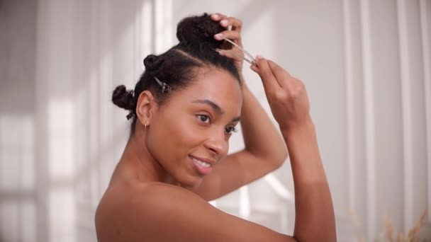 Černá žena dělat účes pro afro vlasy a usmívat se na kameru ve slunném pokoji — Stock video
