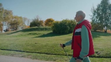 Kadın gülerek yürüyor güneşli sonbahar parkında İskandinav yürüyüş direkleriyle, yan gözle