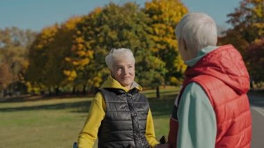 İki yaşlı kadın güneşli sonbahar parkında yelek giyip, baston tutarak konuşuyor.