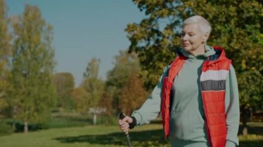 Kadın gülerek yürüyor güneşli sonbahar parkında İskandinav yürüyüş direkleriyle, yan gözle