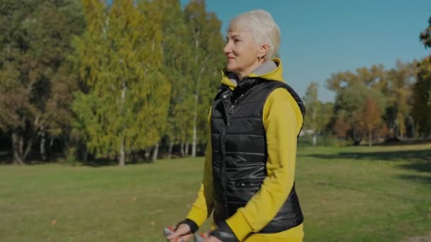 У віці жінка робить скандинавські прогулянки в сонячному осінньому парку самостійно, вид спереду — стокове відео