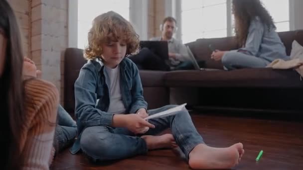 Dzieci robią papierowe samoloty i rysują na podłodze, ich rodzice siedzą na kanapie — Wideo stockowe