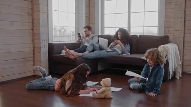 Eltern sitzen auf der Couch und schauen auf Telefone, während Kinder auf dem Boden zeichnen — Stockvideo