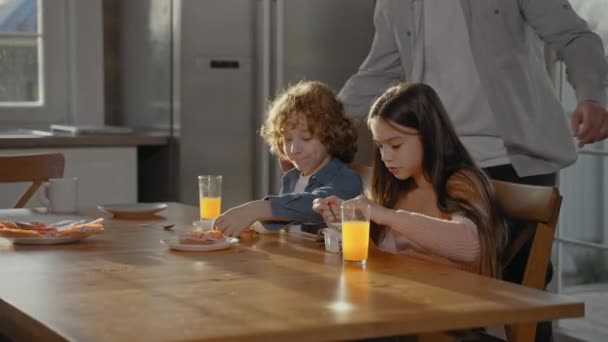 Az ember megöleli a gyerekeit narancslevet iszogatva reggelinél egy napsütéses napon. — Stock videók