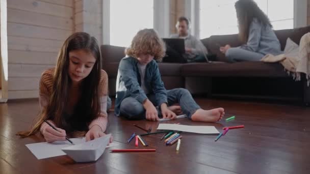 Τα παιδιά φτιάχνουν χάρτινα αεροπλάνα και ζωγραφίζουν στο πάτωμα, οι γονείς τους κάθονται στον καναπέ. — Αρχείο Βίντεο