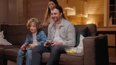 Baba oğluyla oyun konsolunda oynuyor ve kazanıyor, kanepede oturuyor.