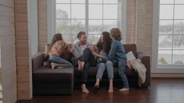 Νεαρή οικογένεια κάθονται μαζί σε έναν καναπέ γαργαλώντας και χαμογελώντας, κοιτάζοντας την κάμερα — Αρχείο Βίντεο