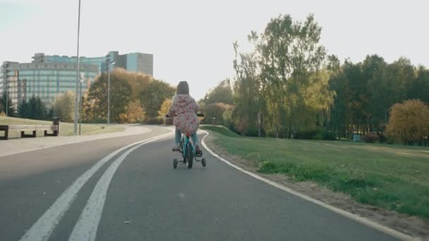 Маленька дівчинка їде на велосипеді в міському парку на заході сонця восени — стокове відео