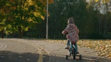 Küçük kız güneşli sonbaharda gün batımında parkta bisiklet sürüyor.