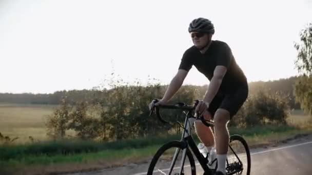 Велосипедист їде на дорожньому велосипеді на заході сонця на шосе біля поля, в шоломі — стокове відео