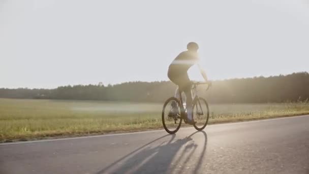 Ciclista em uma bicicleta de estrada ao pôr do sol em uma estrada perto do campo, usando capacete — Vídeo de Stock