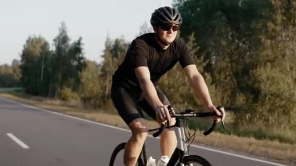 Чоловік їде на дорожньому велосипеді на заході сонця на шосе поблизу поля, в шоломі — стокове відео