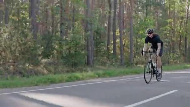 Um ciclista masculino montando uma bicicleta de estrada em uma floresta ensolarada, usando capacete — Vídeo de Stock