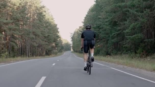 Tiro nas costas de um homem andando de bicicleta de estrada em uma floresta ao pôr do sol, usando capacete — Vídeo de Stock